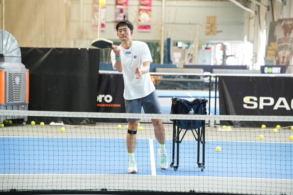 A Sideout School coach with a paddle and basket of balls beside the net